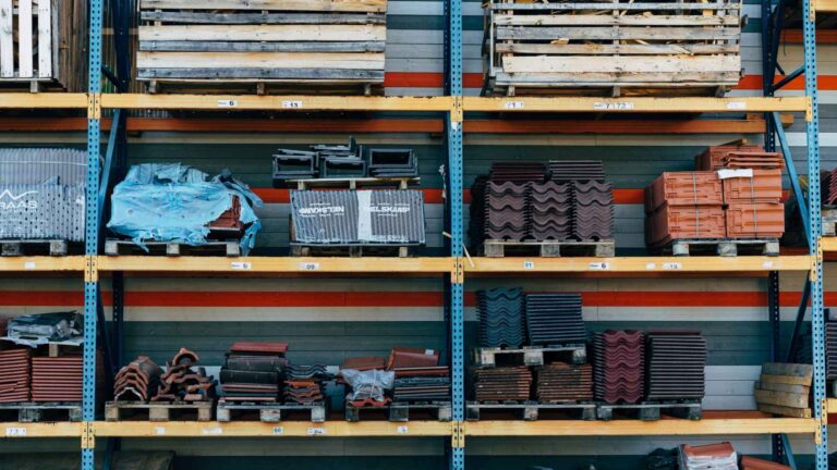 Roofing materials including asphalt shingles, metal panels and tiles stacked on warehouse shelves